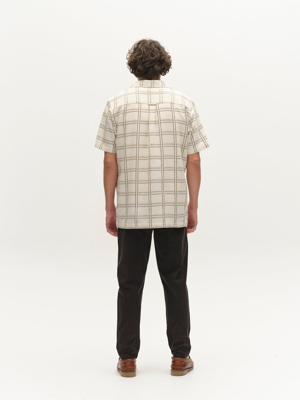 A person with short curly hair wears a short-sleeve beige plaid shirt, Gabba GABmonza Tasca Bolu TENCEL™ Pant in black, and brown shoes, standing facing away against a plain white background.