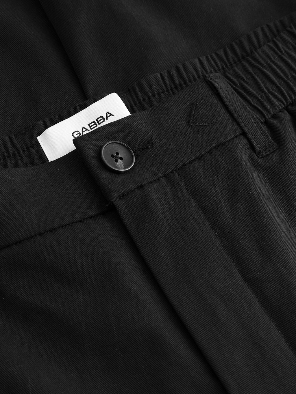 Close-up of the GABmonza Tasca Bolu TENCEL™ Pant in black by Gabba, showing relaxed straight fit, elastic waistband, black button, and a white label with the brand name Gabba partially visible.