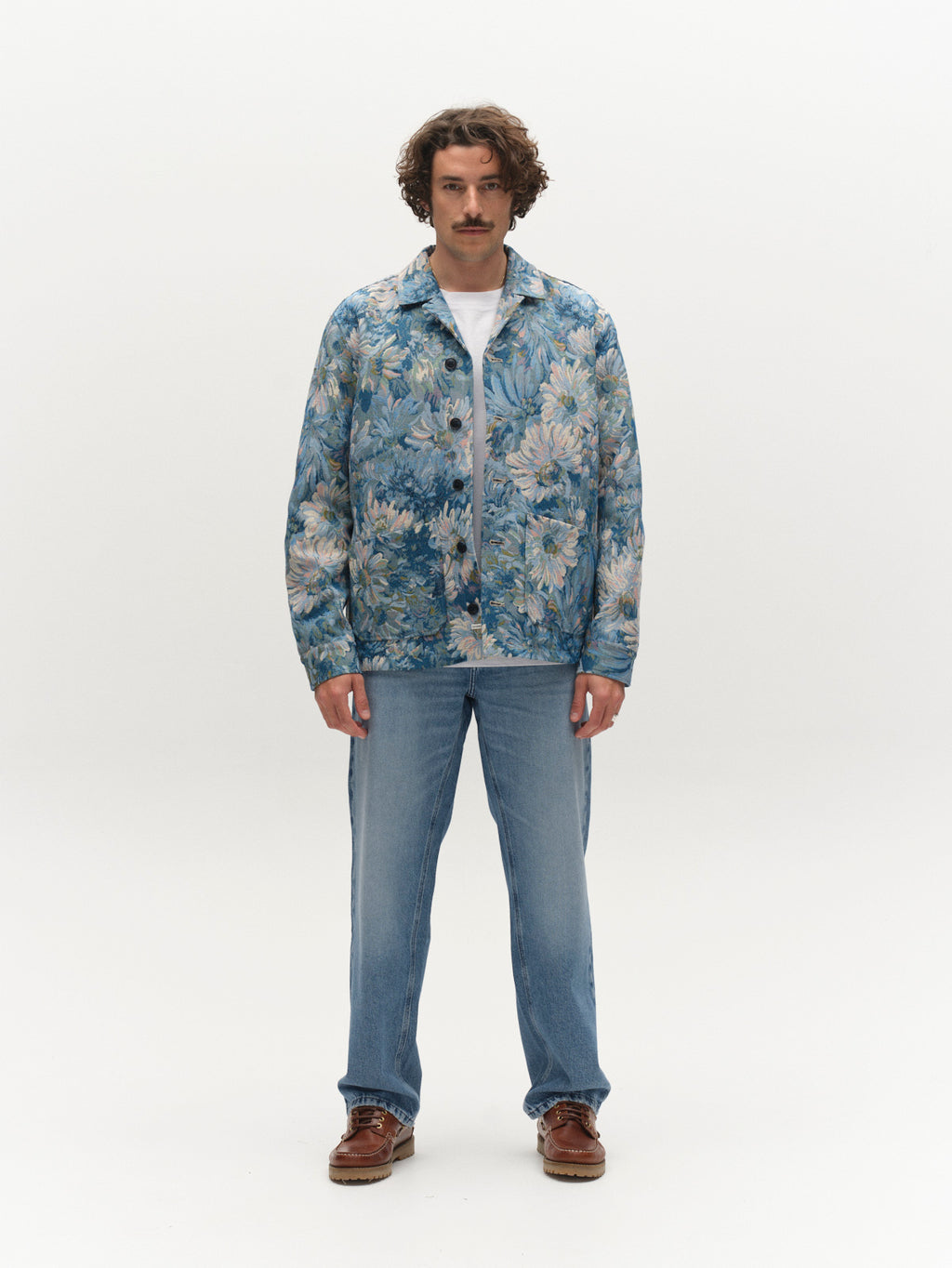A man with curly hair and a mustache stands facing forward, wearing the Gabba GABsimone Art LS - Blue Pattern overshirt over a white shirt, light blue jeans, and brown shoes against a plain white background.