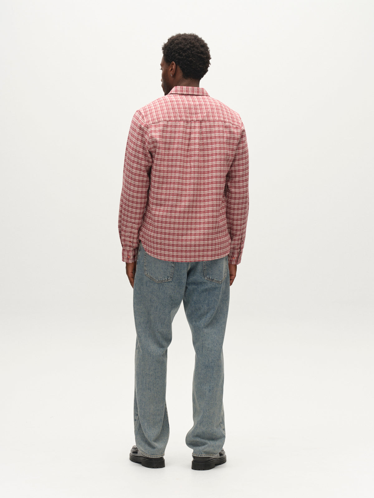 A man stands facing away from the camera, wearing the Gabba GABsven Reg TX2 LS Shirt in Brick Red—a regular-fit, yarn-dyed cotton shirt with a checked texture—paired with light blue jeans and black shoes, against a plain white background.