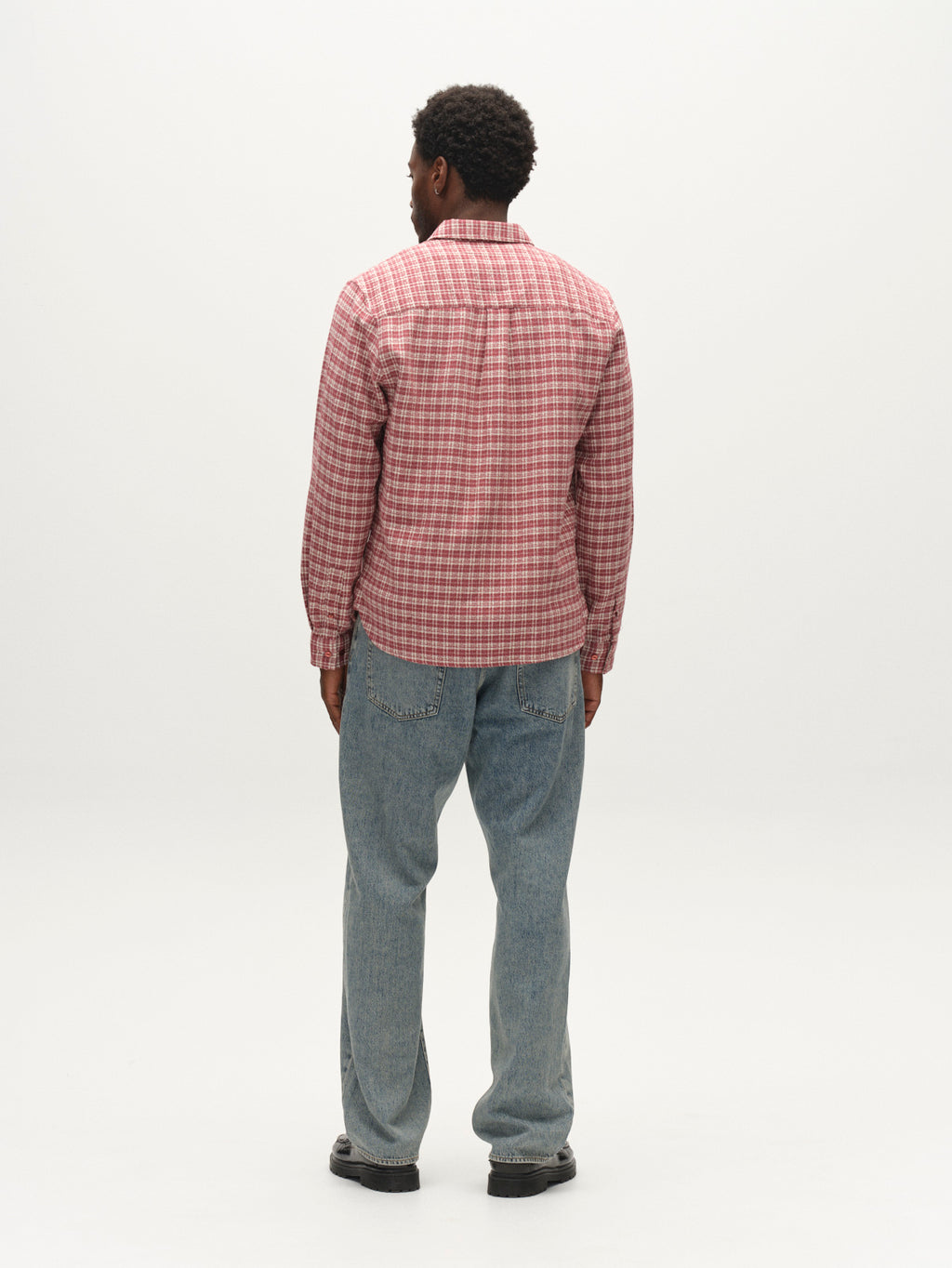A man stands facing away from the camera, wearing the Gabba GABsven Reg TX2 LS Shirt in Brick Red—a regular-fit, yarn-dyed cotton shirt with a checked texture—paired with light blue jeans and black shoes, against a plain white background.