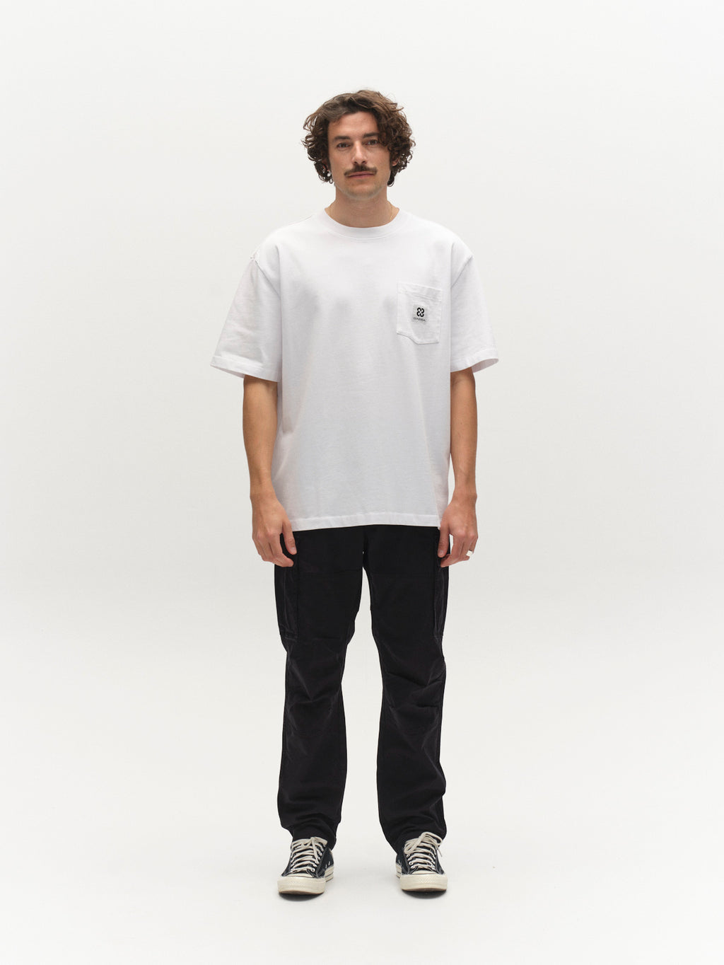 A man with curly hair and a mustache faces forward against a white background, wearing the Gabba GABnigel Wix Pkt Boxy Hour SS T-Shirt in white, paired with black pants and black-and-white sneakers.