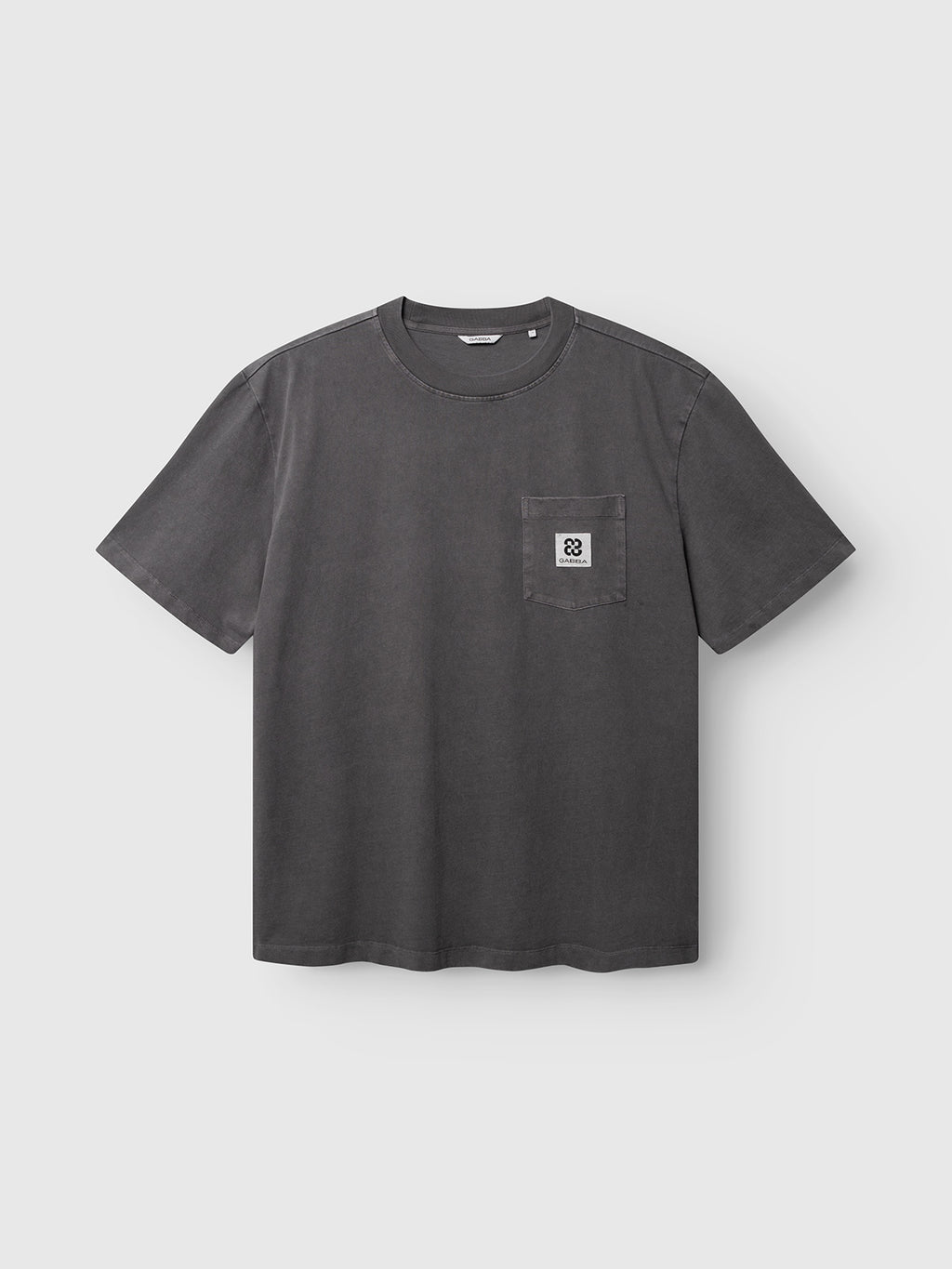 The Gabba GABnigel Wix Pkt Boxy Hour SS T-Shirt in dark grey is a heavyweight cotton tee with short sleeves, a round neck, a left chest pocket and a white patch with black geometric design.