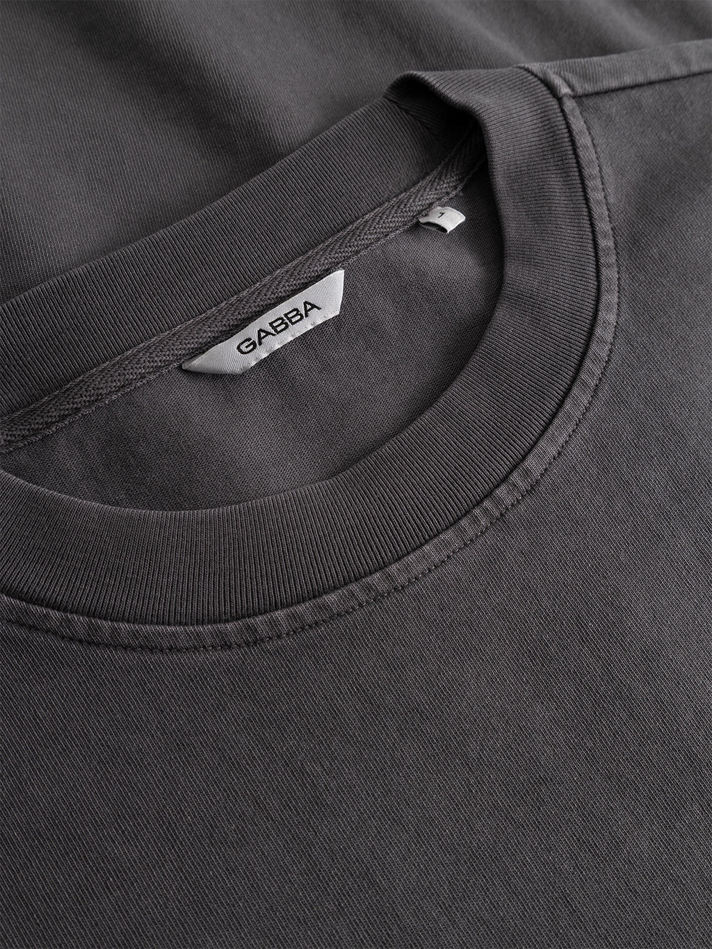 Close-up of the Gabba GABnigel Wix Pkt Boxy Hour SS T-Shirt in dark grey, highlighting the collar and neckline. The inside label shows 