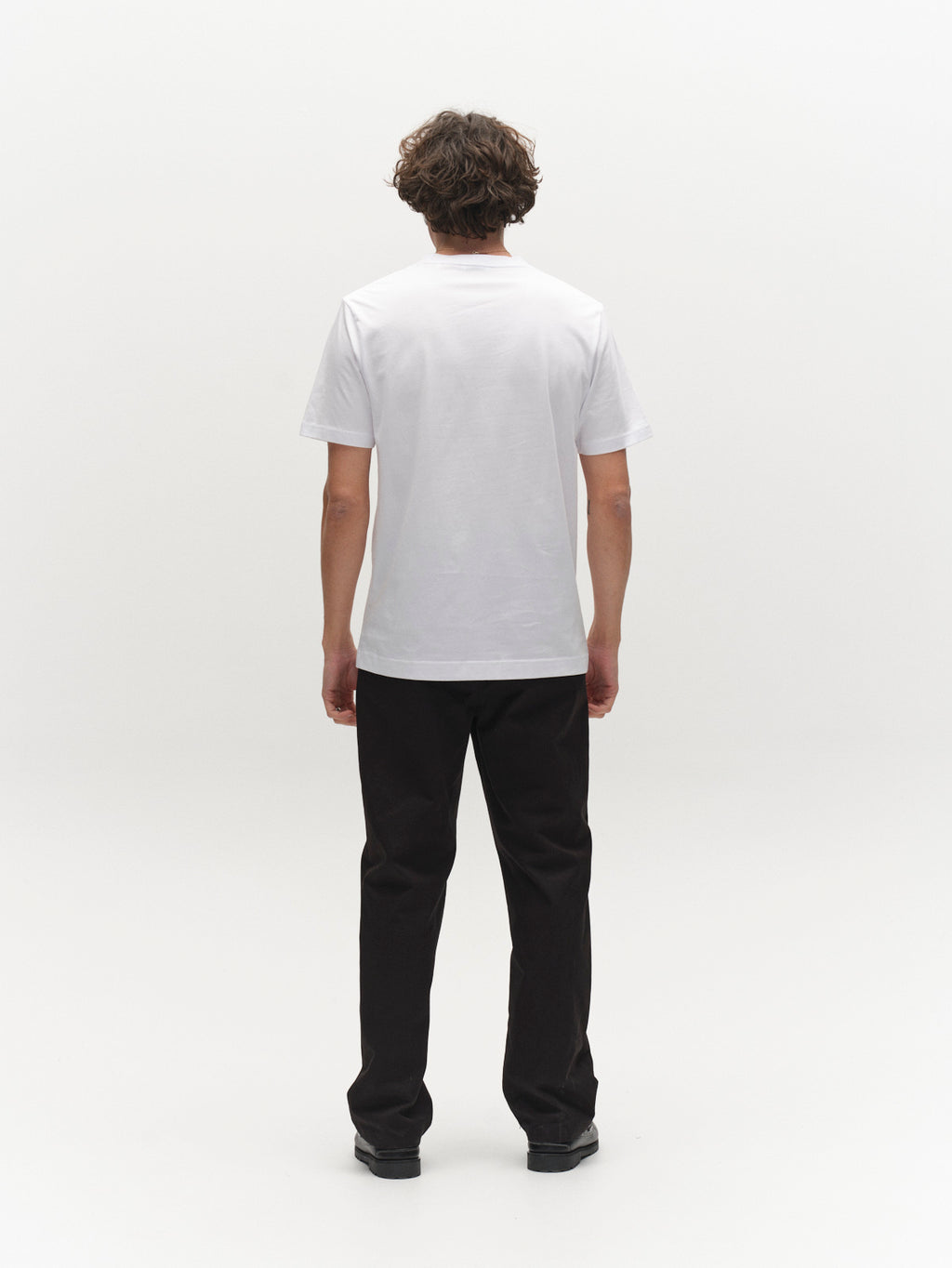 A person with short, curly hair stands facing away, wearing the Gabba GABdune Element GOTS SS T-Shirt in white, paired with black pants and black shoes against a plain white background.