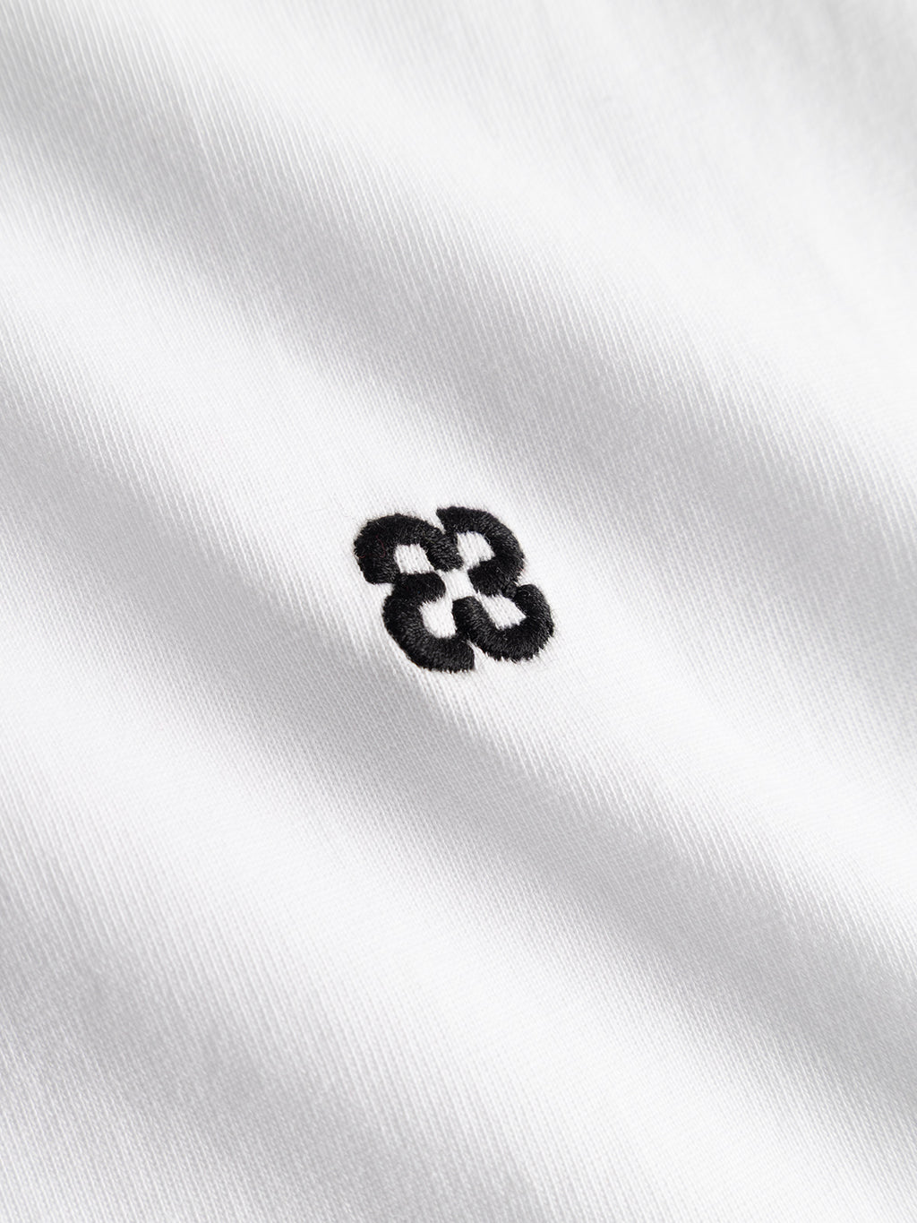 Close-up of the Gabba GABdune Element GOTS SS T-Shirt in white, a classic fit made from organic cotton. Features a small black geometric embroidery with four interlocking loops at the center; texture and stitching are visible.