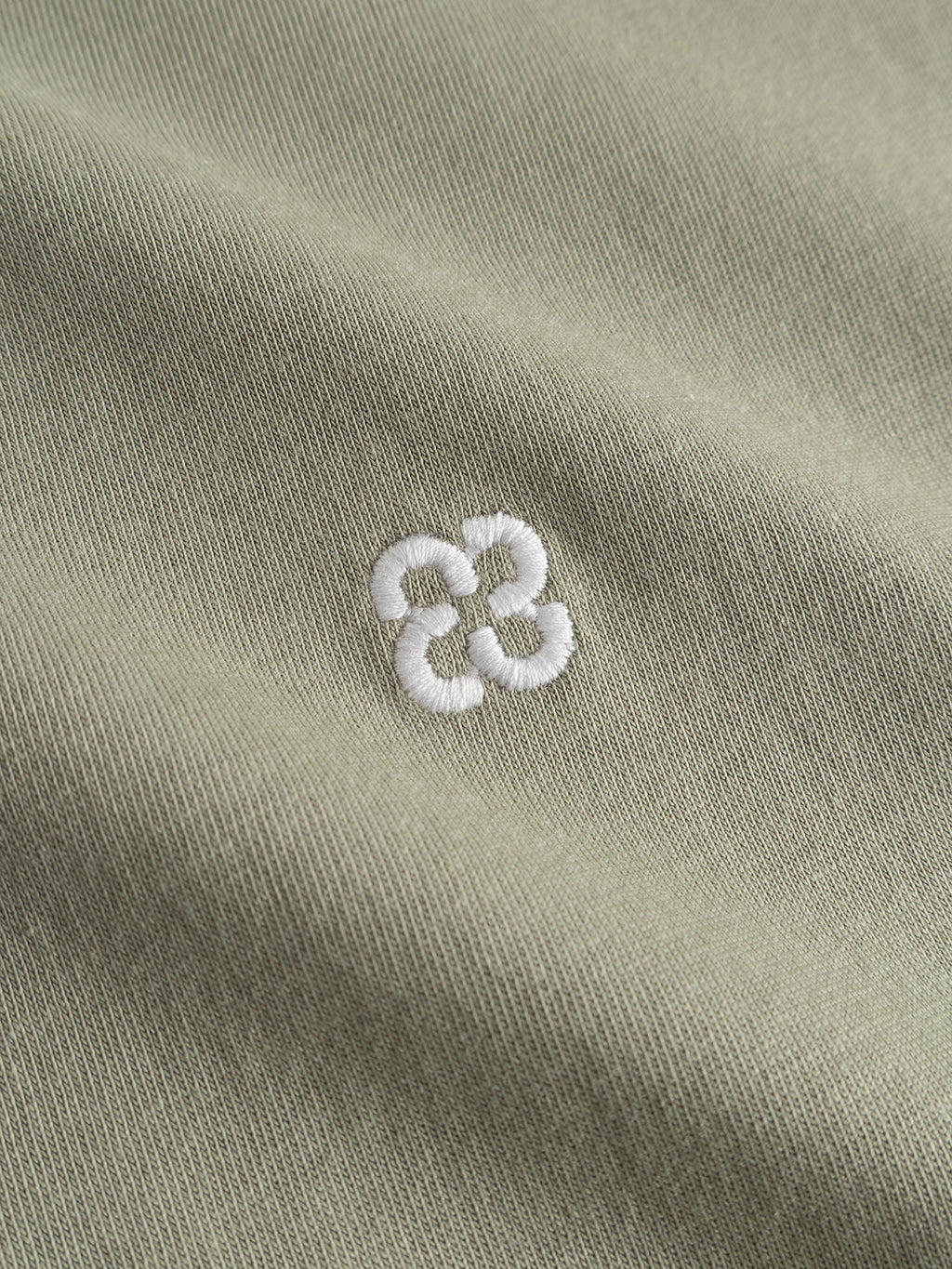 Close-up of Gabba's GABdune Element GOTS SS T-Shirt in light green, featuring soft, light olive organic cotton fabric with a small white embroidered flower-like design at the center. Made from GOTS-certified material with gentle folds.