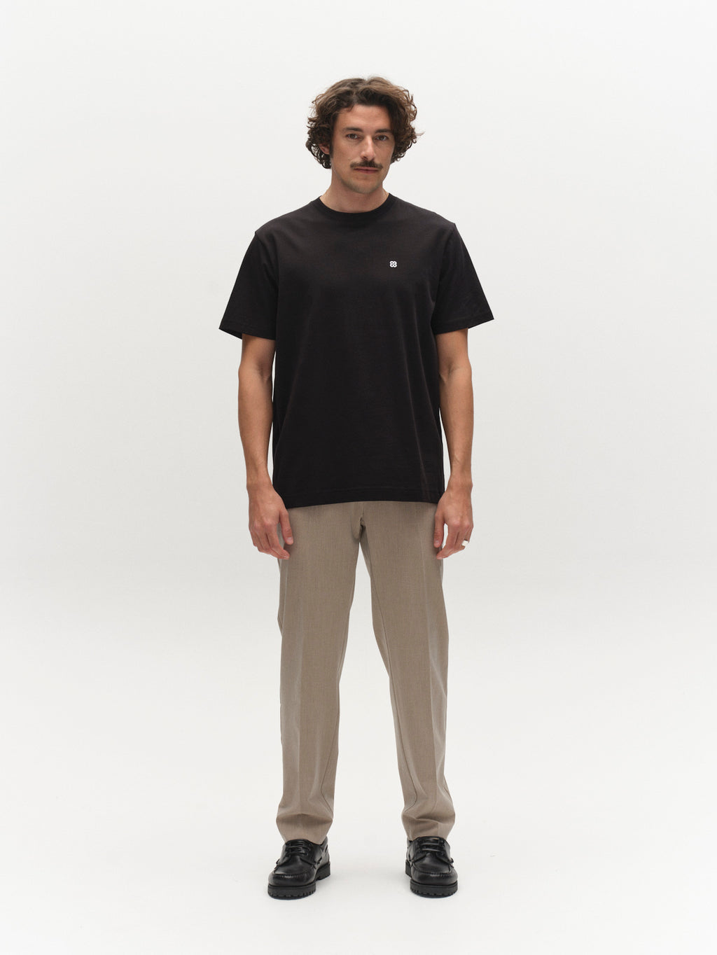 A man with wavy brown hair and a mustache stands facing forward against a plain white background, wearing a Gabba GABdune Element GOTS SS T-Shirt in black, beige pants, and black shoes.