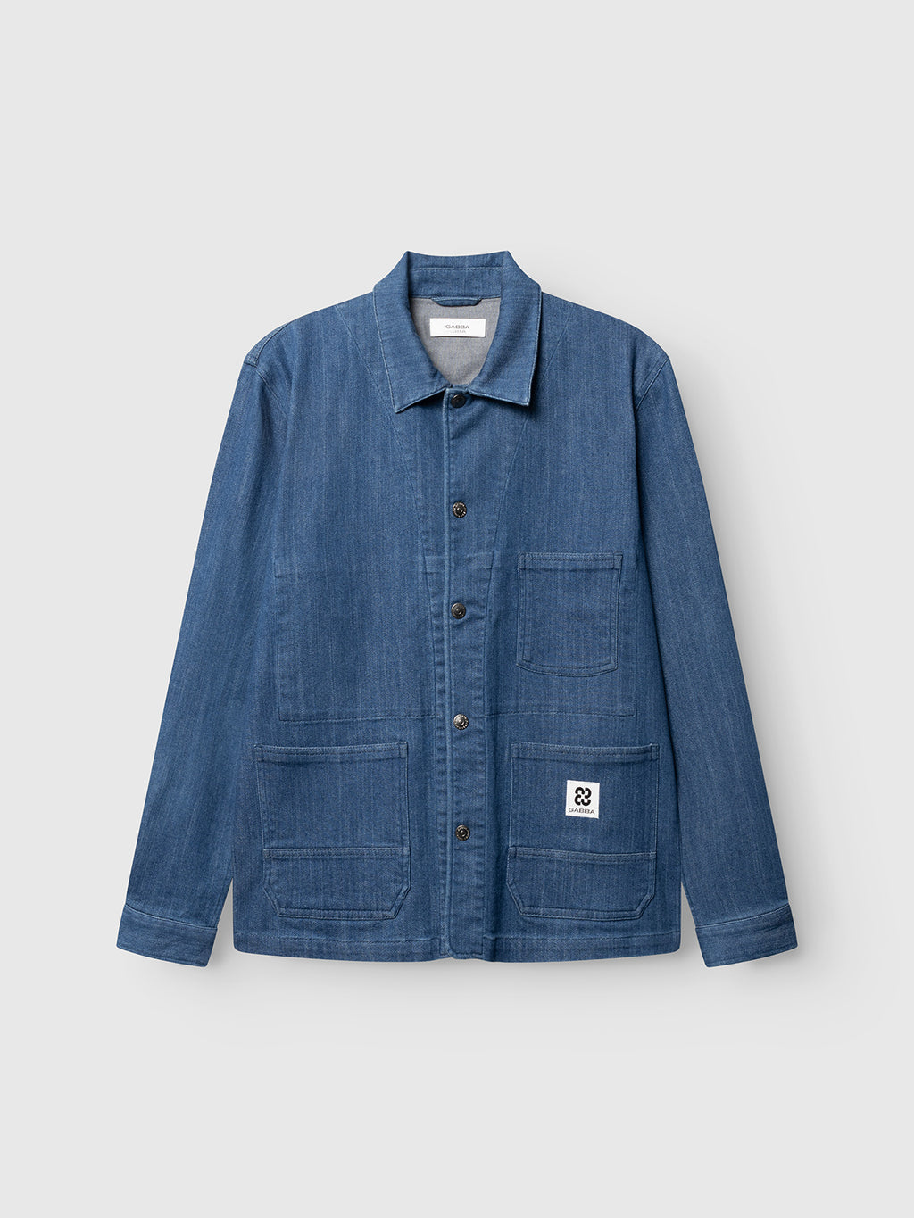 The Gabba GABbeta Denim LS Overshirt in mid blue denim is made from stretch cotton, featuring a collar, button front, long sleeves, and four pockets—one with a white patch and black logo. Shown against a plain light background.