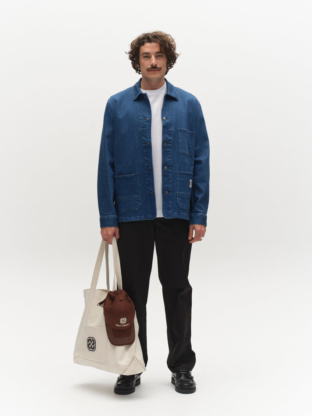 A man with wavy hair and a mustache faces forward, wearing the Gabba GABbeta Denim LS Overshirt in Mid Blue Denim over a white shirt, with black pants, black shoes, and holding a beige tote bag containing a brown drawstring pouch.