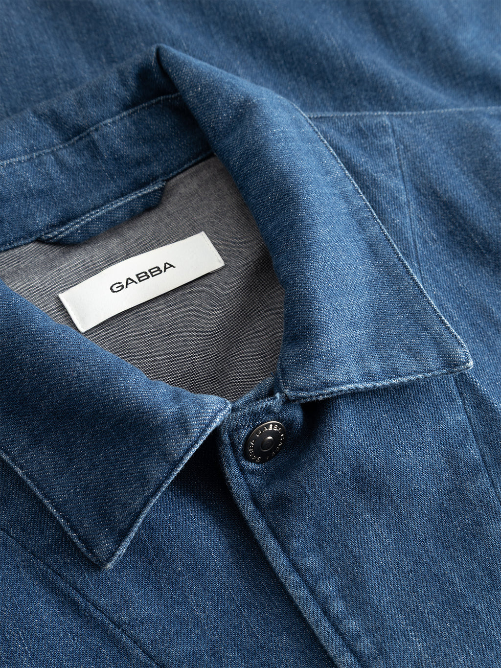 Close-up of the Gabba GABbeta Denim LS Overshirt in mid blue denim, featuring a stretch cotton collar with black button and white Gabba label inside, laid flat to highlight its durable workwear fabric and classic design.
