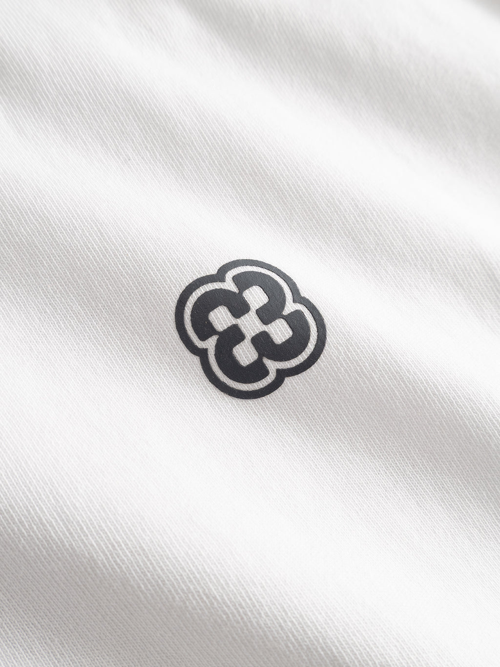 A close-up of the Gabba GABlegin Wix Chap Boxy Hour SS T-Shirt in white, featuring soft, heavyweight cotton fabric with a small black geometric floral logo printed at the center.