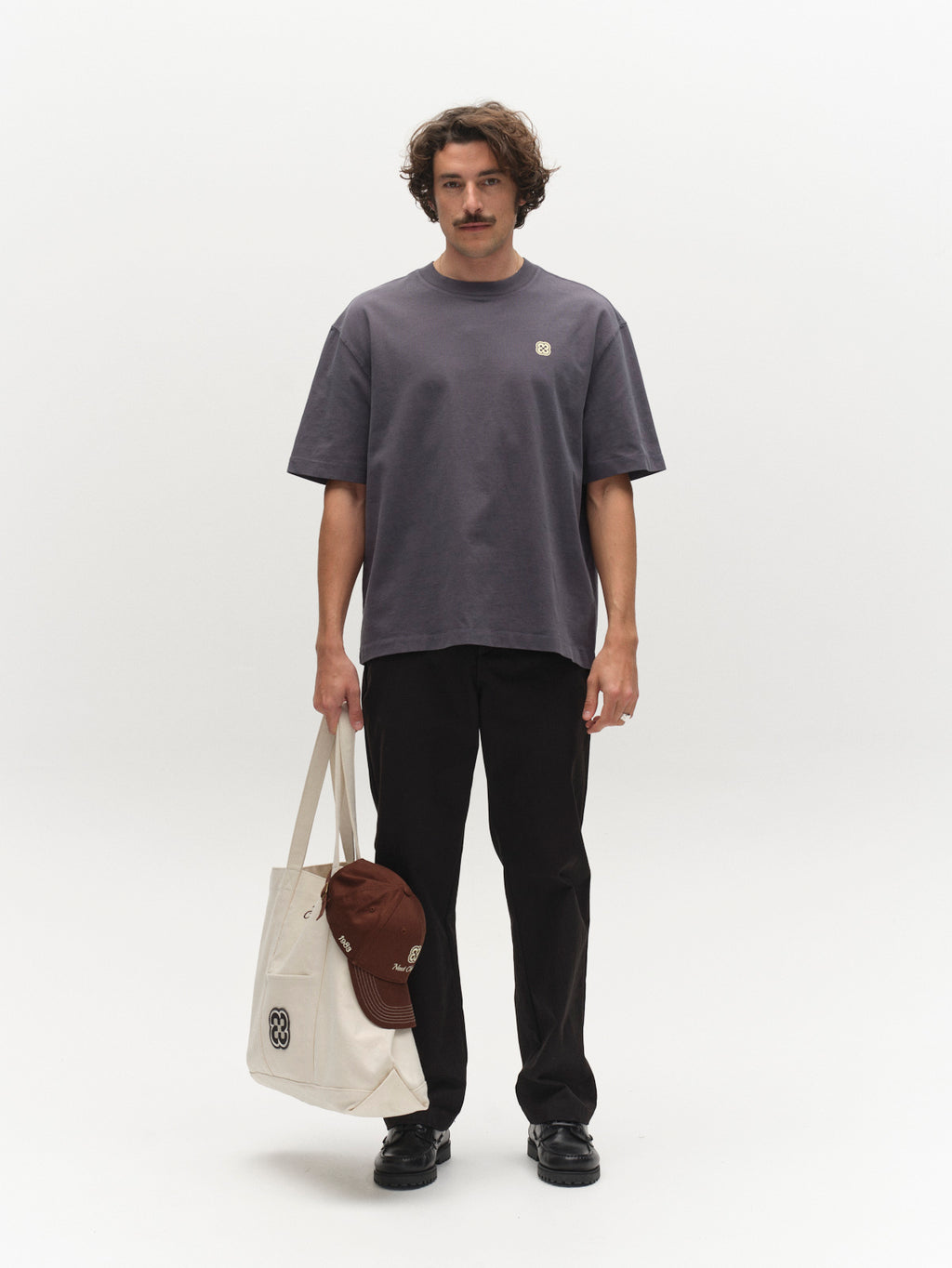 A man with wavy hair and a mustache stands against a white background, wearing the Gabba GABlegin Wix Chap Boxy Hour SS T-Shirt in Turbulence Blue, paired with black pants, black shoes, and holding a cream tote bag with a brown cap attached.