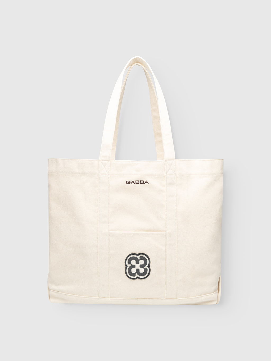 The Gabba GABchap Tote Bag in off-white has two handles and a front pocket with “GABBA” in black above an embroidered black and white flower logo. The sturdy canvas bag is shown against a plain light gray background.