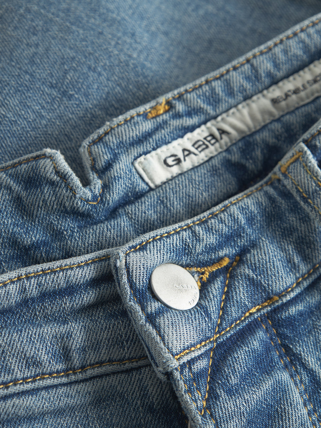 Close-up of Gabba GABalex Cros Cropped Lt. Blue Denim jeans with a silver button, yellow stitching, and a visible white label featuring the Gabba brand name inside the waistband.