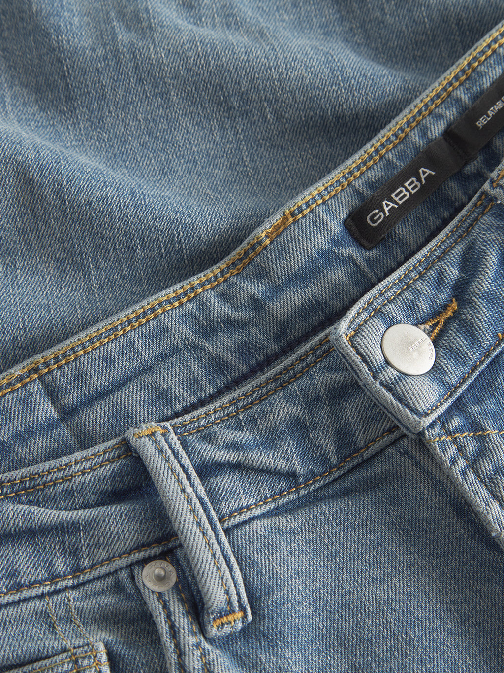 Close-up of GABmax Cros - Lt. Blue Denim jeans by Gabba, featuring a sharp modern slim fit, visible fabric texture and stitching, silver button, waistband, belt loops, and a sleek black brand label.