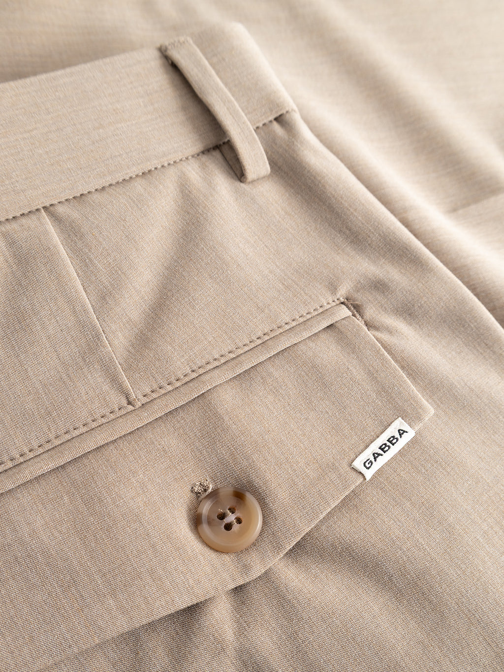 Close-up of GABjoli Tailor Dressy Shorts in sand by Gabba, highlighting the beige fabric, a back pocket with button, white Gabba label, and a visible belt loop.