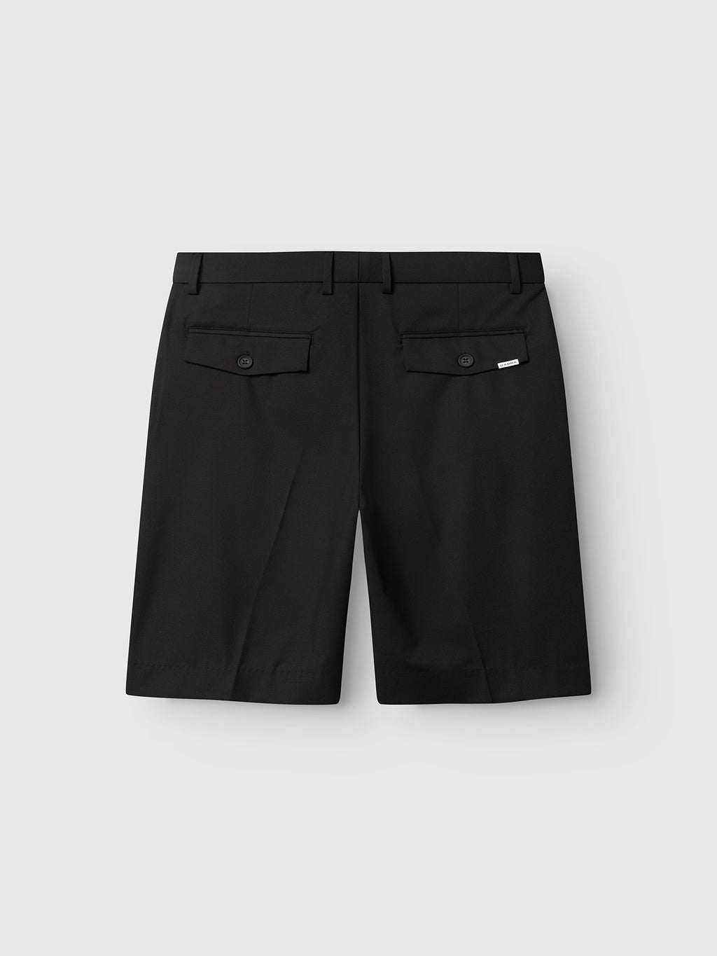 GABjoli Tailor Dressy Shorts by Gabba in black, knee-length with a relaxed fit, two buttoned flap back pockets, and belt loops. Displayed on a plain light gray background with a subtle Gabba brand mark accentuating the tailored style.