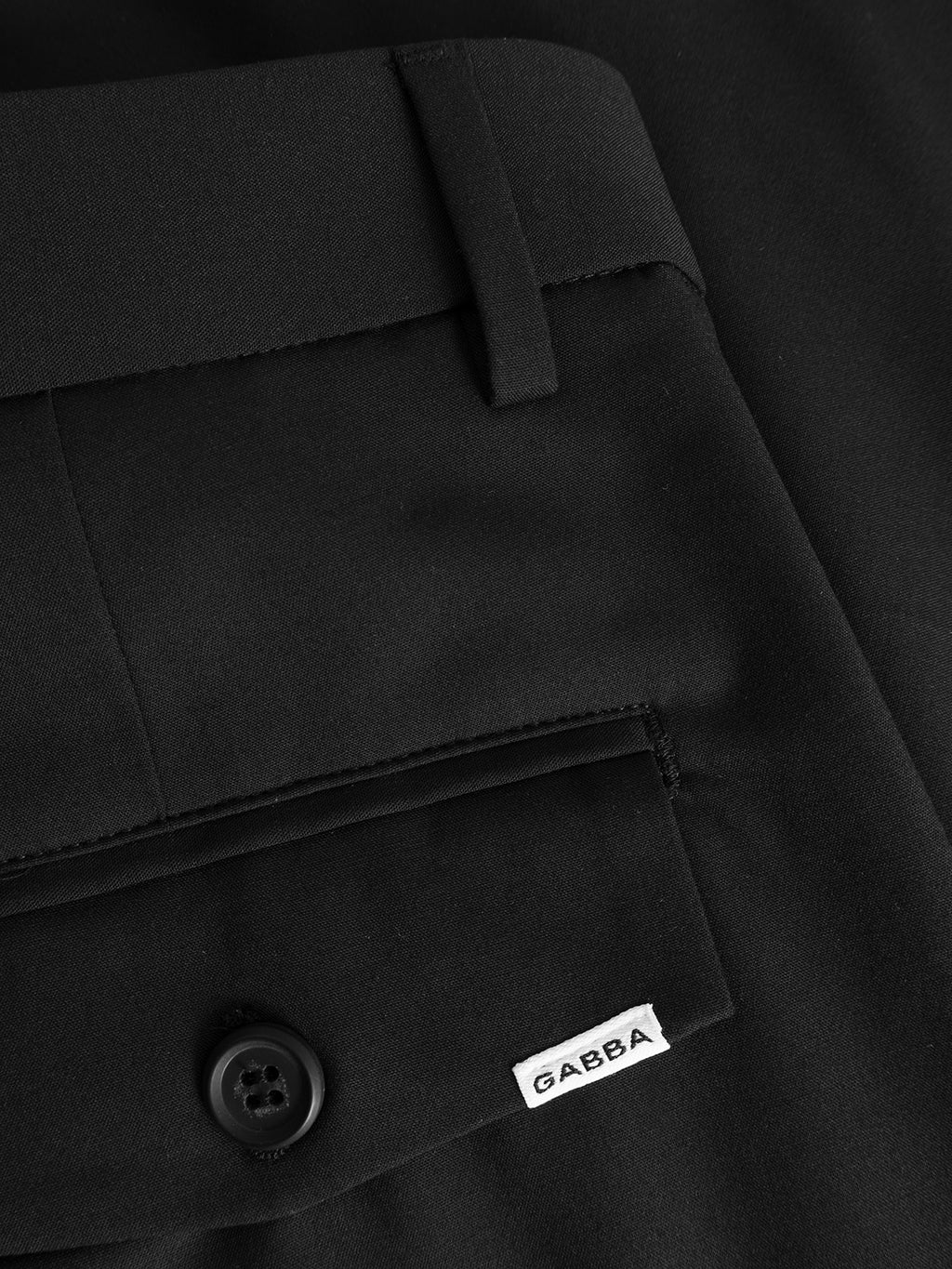 Close-up of GABjoli Tailor Dressy Shorts in black by Gabba, featuring a pocket with a black button and a white fabric tag showing the Gabba logo. The tailored design highlights detailed stitching and rich fabric texture.