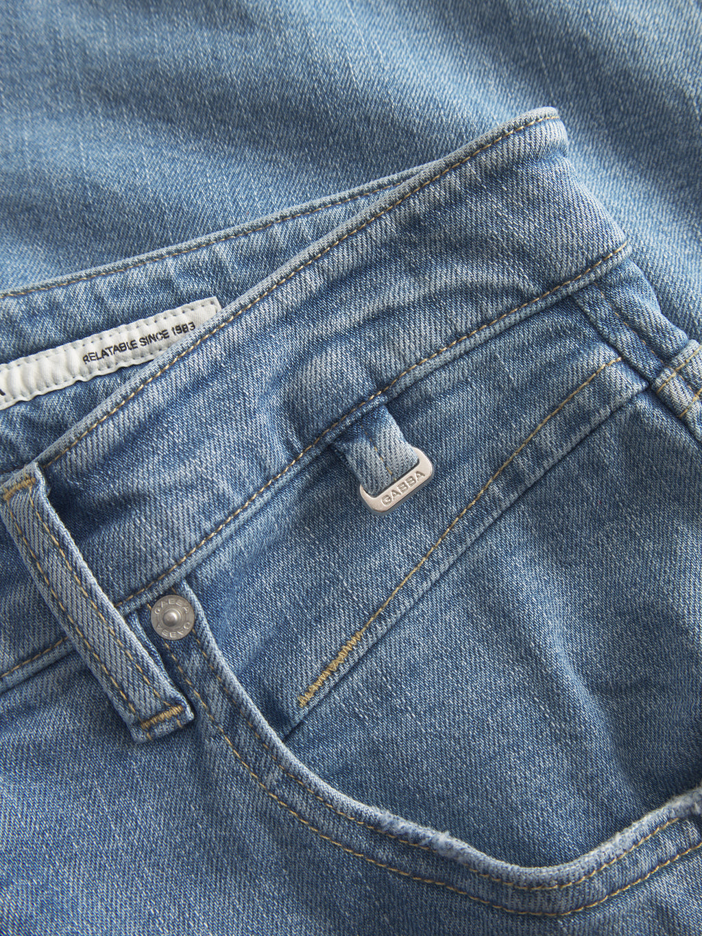 Close-up of GABalex Cross - Lt. Blue Denim jeans by Gabba, showing the front pocket, waistband, visible button, and a small white tag. The jeans are laid flat with the denim fabric texture clearly visible.