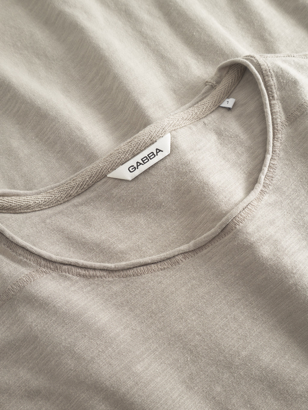 A close-up of the GABkostant New SS t-shirt by Gabba in Grey Paloma, featuring light beige slub fabric, a visible brand tag at the neckline, unique texture, 100% cotton composition, and detailed stitching.
