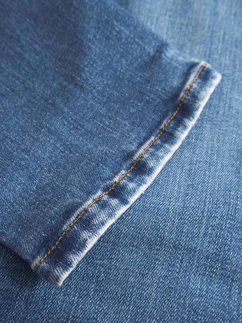 Close-up of the hem of a blue denim pant leg, detailed with orange stitching and a faded edge, lying atop another piece of blue denim fabric—these Gabba GABjones2 Nham RS2113 Jeans are crafted from dark blue performance stretch denim in a slim straight fit, offering both style and comfort.