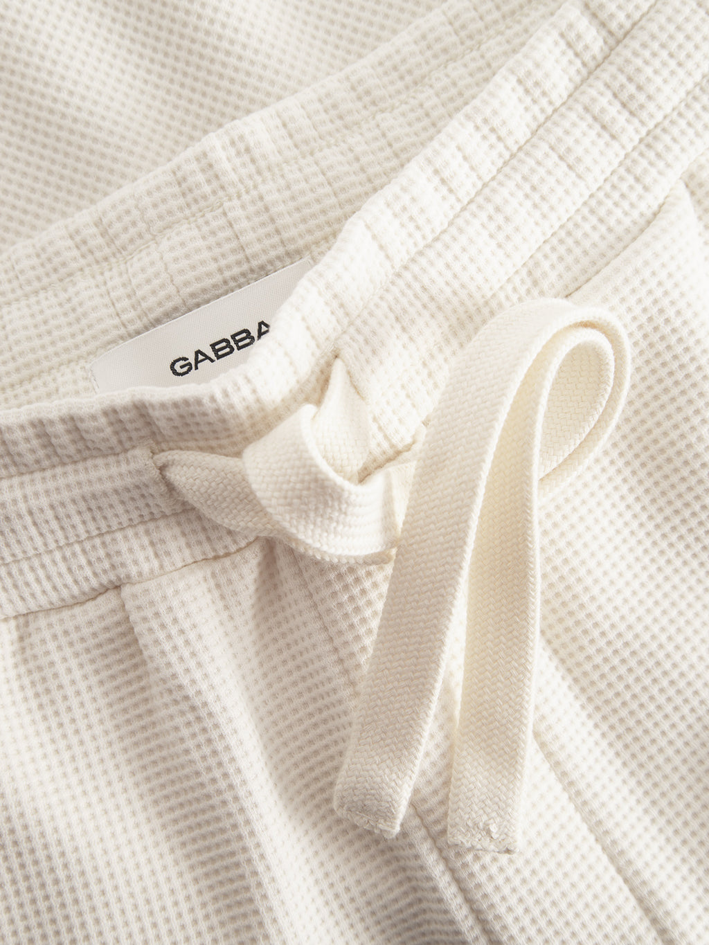 Close-up of Tim Waff - Bone White pants by Gabba, featuring white textured fabric, drawstring waistband, tie detail, and a partially visible Gabba label.