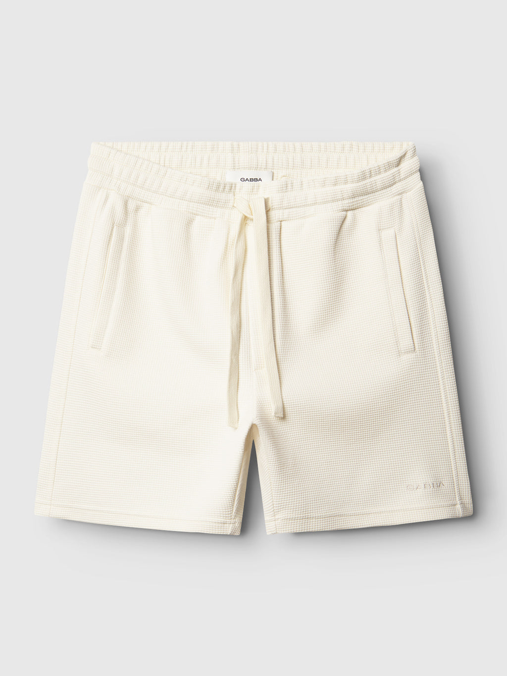 Gabba Tim Waff - Bone White shorts feature a cream waffle-knit design with elastic waistband, drawstring, side pockets, and a subtle logo on the lower left leg, shown flat on a light background.