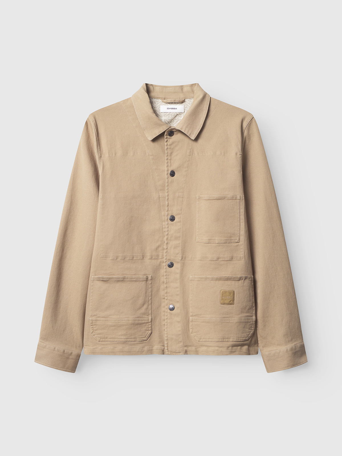 GABbeta Twill - Sand by Gabba is a beige canvas chore jacket featuring a collar, button front, long sleeves, and three front utility pockets. Made from garment-dyed stretch twill for enhanced comfort, this regular fit style includes a small square logo patch on the lower right pocket. Photographed against a plain white background.