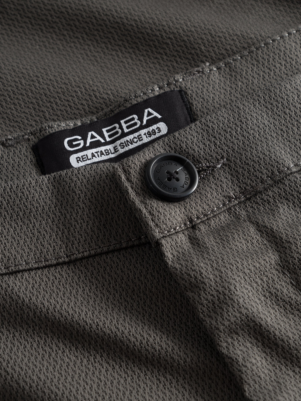 Close-up of Pisa K3280 Dale Pants by GABBA in grey, showing a black button, textured fabric, tapered fit, and a black waistband label with 