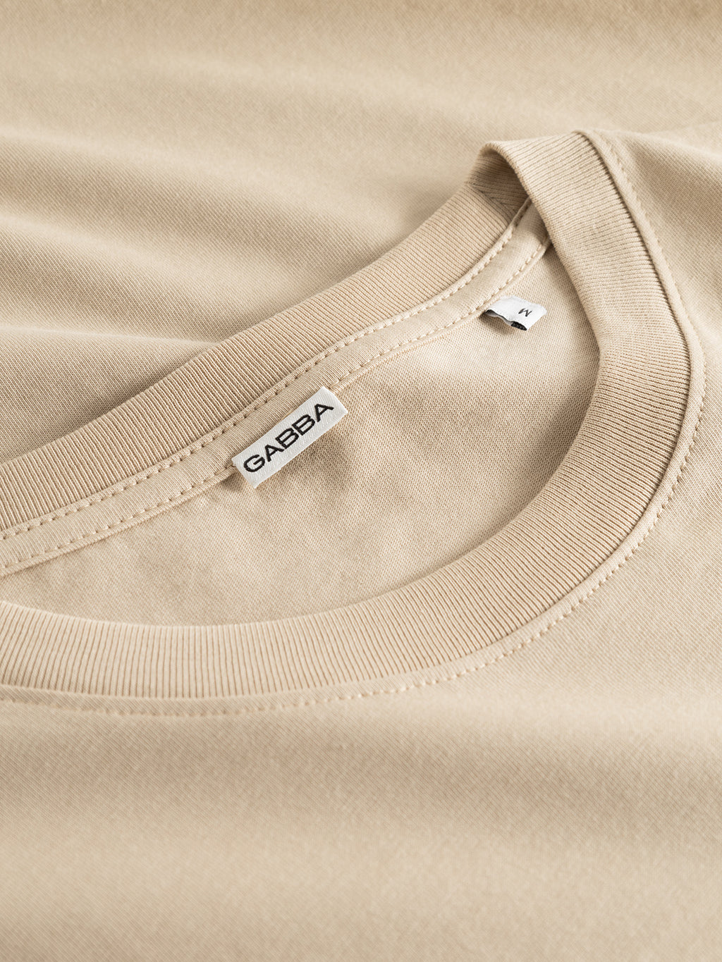 Close-up of the Gabba GABdune Logo GOTS SS T-Shirt in Safari beige, crafted from organic cotton, featuring a visible neckline label, displayed on a matching fabric background.