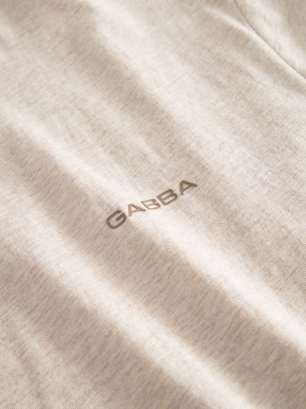 A close-up of Dune Melange SS - Lt. Sand jersey fabric by Gabba, featuring the brand's chest logo in small gray capitals. The beige melange material shows a subtle texture and feels soft to the touch.