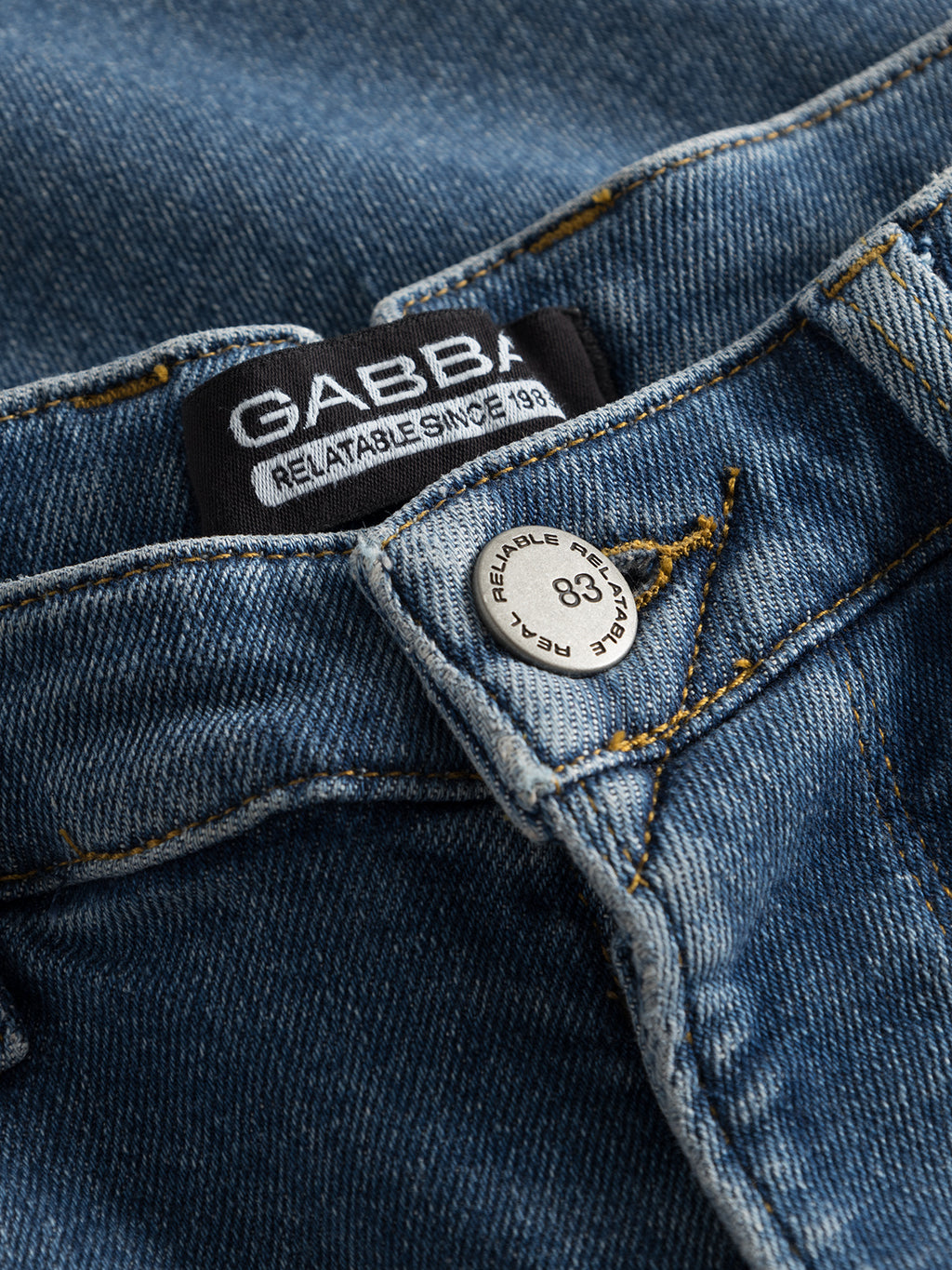 Close-up of GABBA Alex K3948 Jeans in denim wash, featuring a silver button engraved with RELIABLE BRAND 83, a black label with GABBA text and RELIABLE SINCE 1983, plus yellow stitching details.