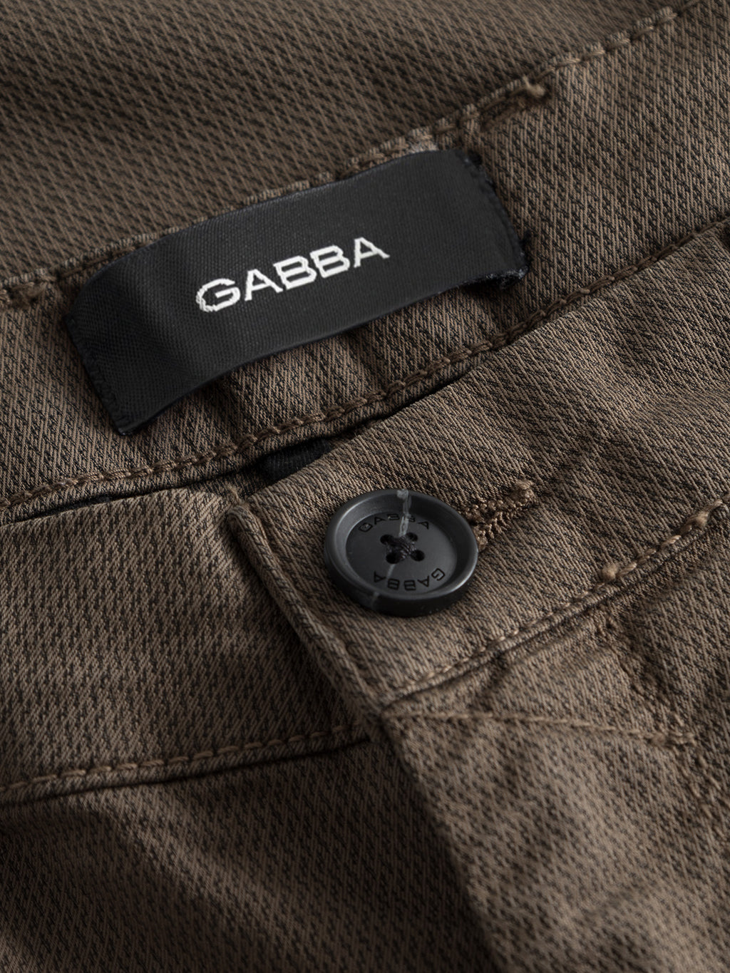 Close-up of GABpaul K3280 Dale Chino - Crocodile by Gabba: brown textured slim chinos with French pockets, a black button, and a black clothing label featuring the Gabba brand name in white text.