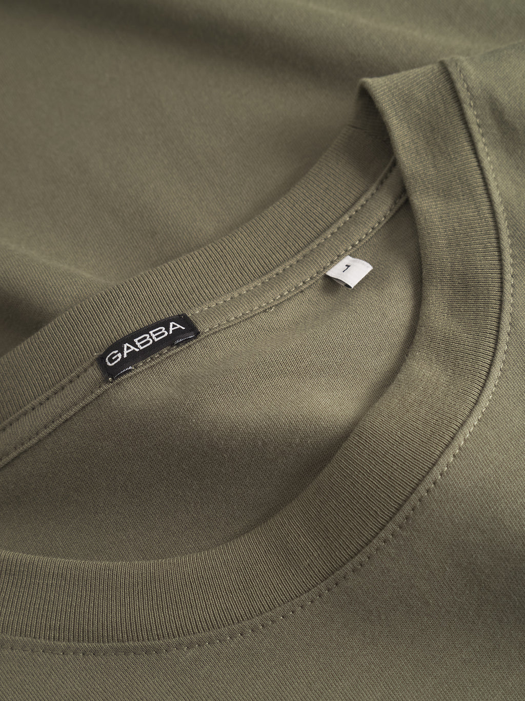Close-up of the Deep Lichen Green GABdune Logo GOTS SS T-Shirt by Gabba, highlighting the classic fit collar and fabric texture. Near the neckline, a black Gabba label and a white size M tag are visible.