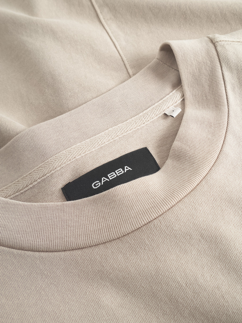 Close-up of the Gabba GABnigel Wix Boxy Hour SS t-shirt in Sand Cashmere, made from heavyweight cotton, featuring a black inner collar label, displayed over matching fabric.