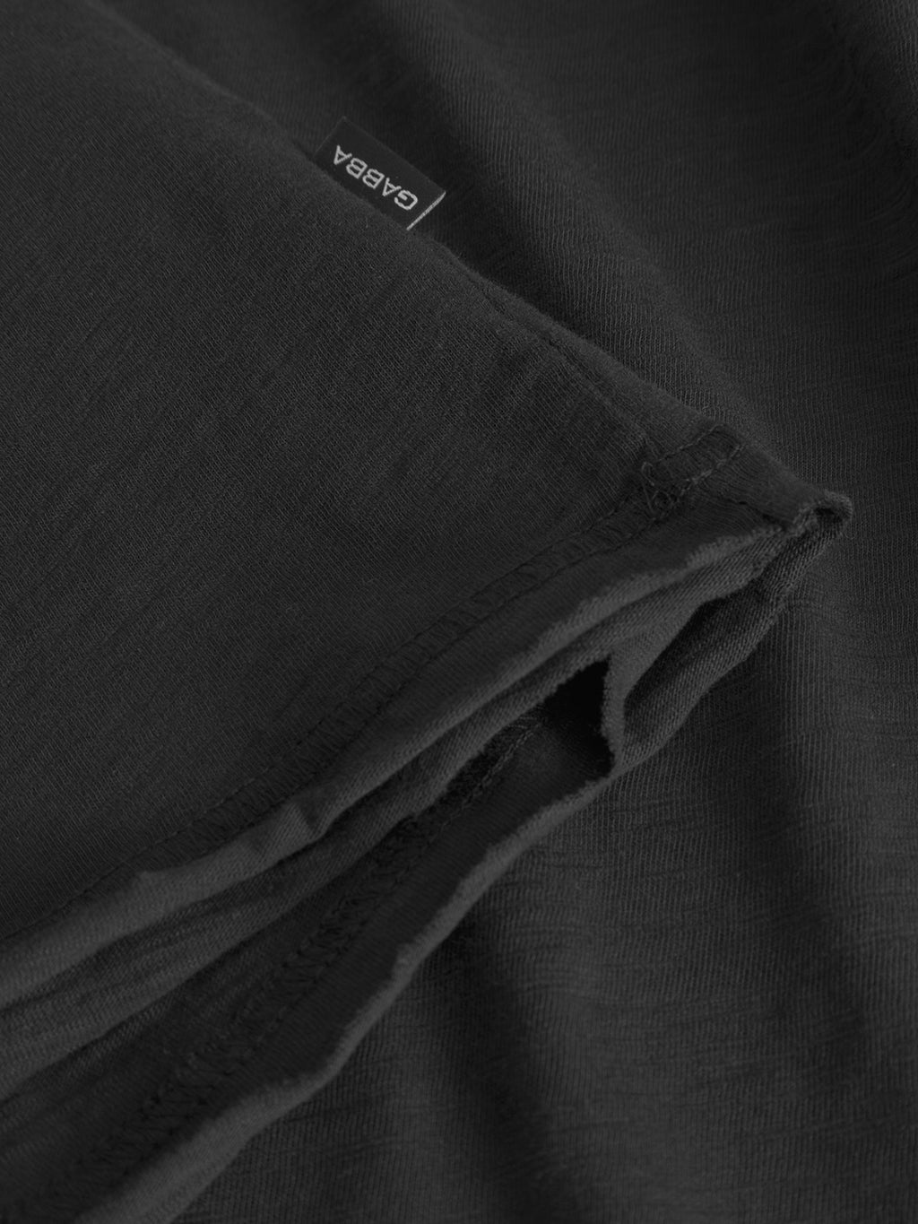Close-up of the Gabba GABkostant New SS - Meteorite t-shirt in black slub fabric, highlighting the hem, a white tag with Gabba branding, and visible textured stitching details.