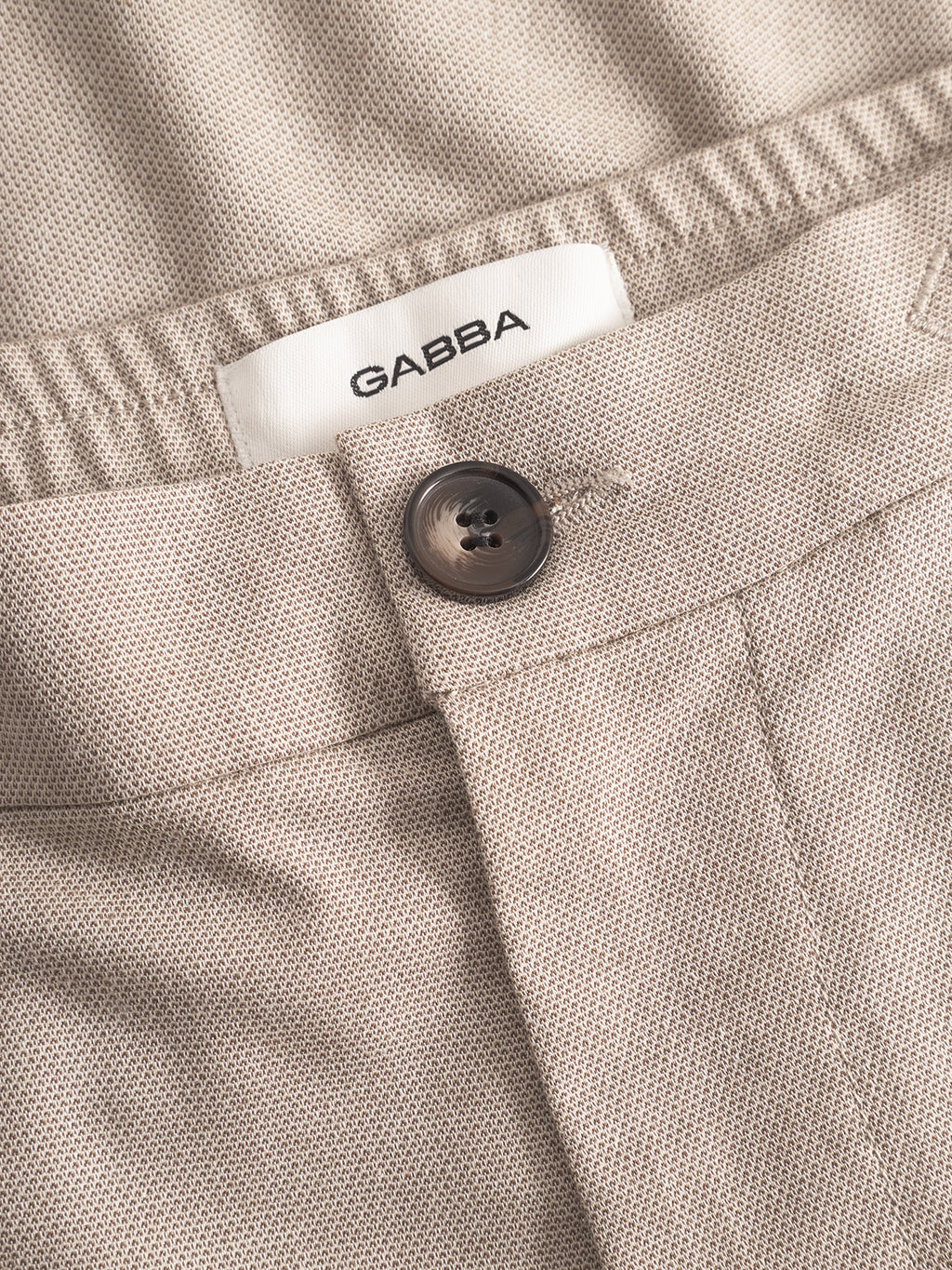 Close-up of Monza Button Carre - Sand relaxed straight pants by Gabba, featuring beige textured fabric, an elastic waistband, single black button, and a white inner label with the brand name—a versatile everyday comfort style.