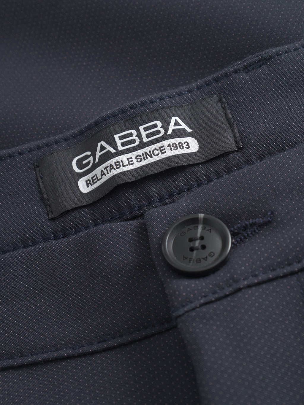 Close-up of a dark fabric label reading GABBA RELATABLE SINCE 1983 next to a matching black button—signature details of the GABBA Pisa Small Dot - Navy pants in high-stretch cotton-blend for modern metro style.