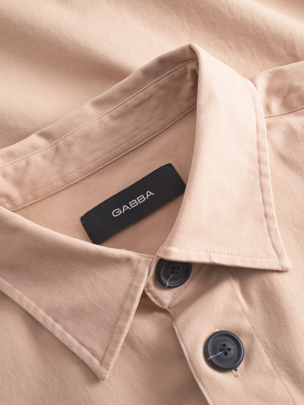 A close-up of the GABBA GABtopper LS Shirt in Humus shows its crisp collar and black buttons. The smooth, neatly pressed fabric and visible brand label give this beige overshirt a polished look.