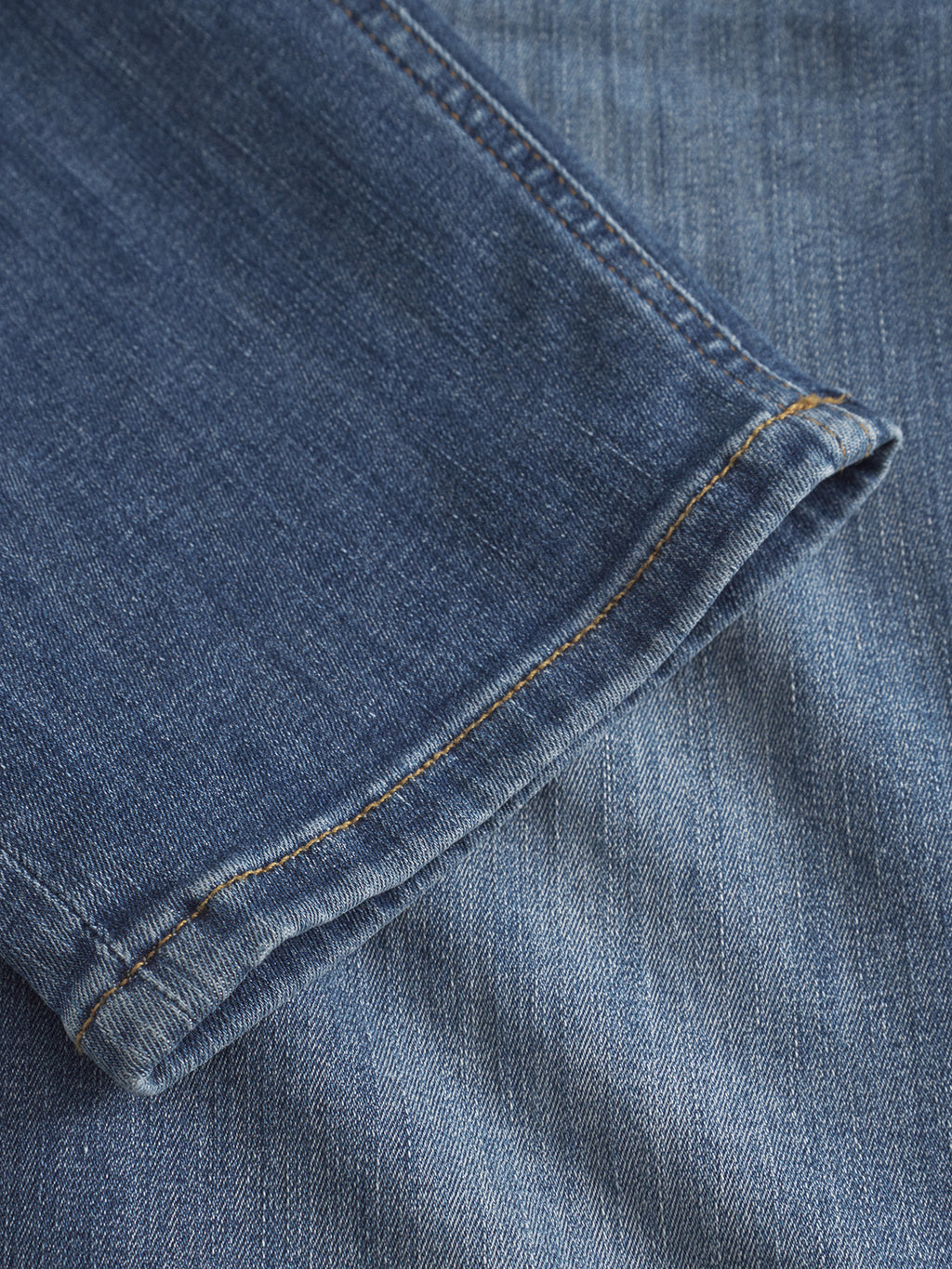 Close-up of the hem of Gabba’s GABjones Vanew - Mid Blue Denim pants, made from high flexibility super-stretch fabric with orange stitching, resting atop another denim piece and highlighting texture and weave in crisp detail.