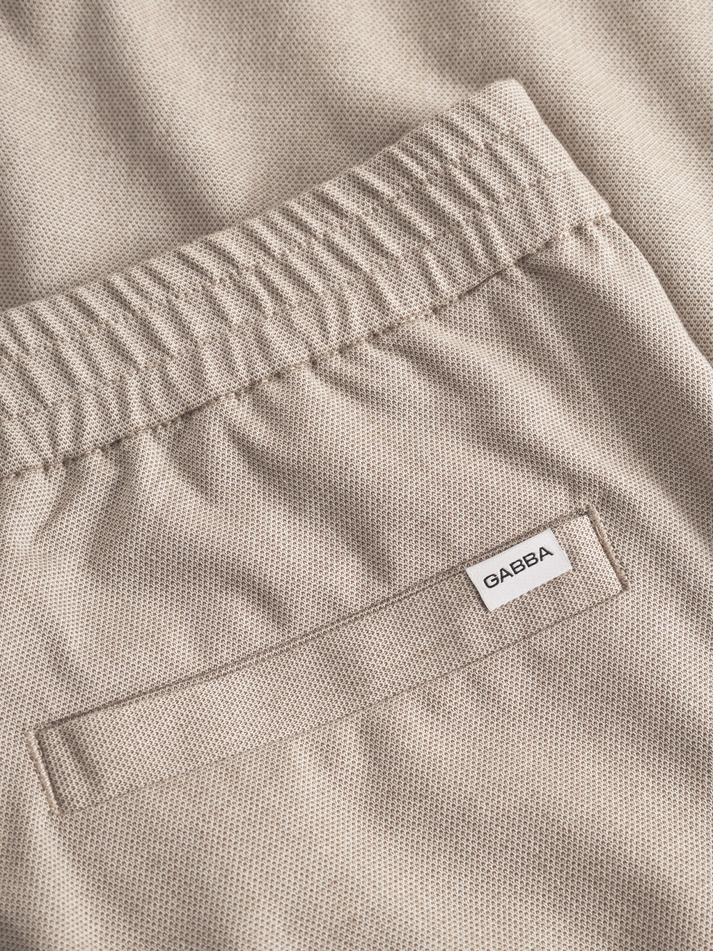 Close-up of the back pocket and elastic waistband of Gabba’s Napoli 19 Carre - Sand pants, featuring a textured beige finish and white Gabba label above the pocket. These classic, comfortable pants are ideal for everyday wear.