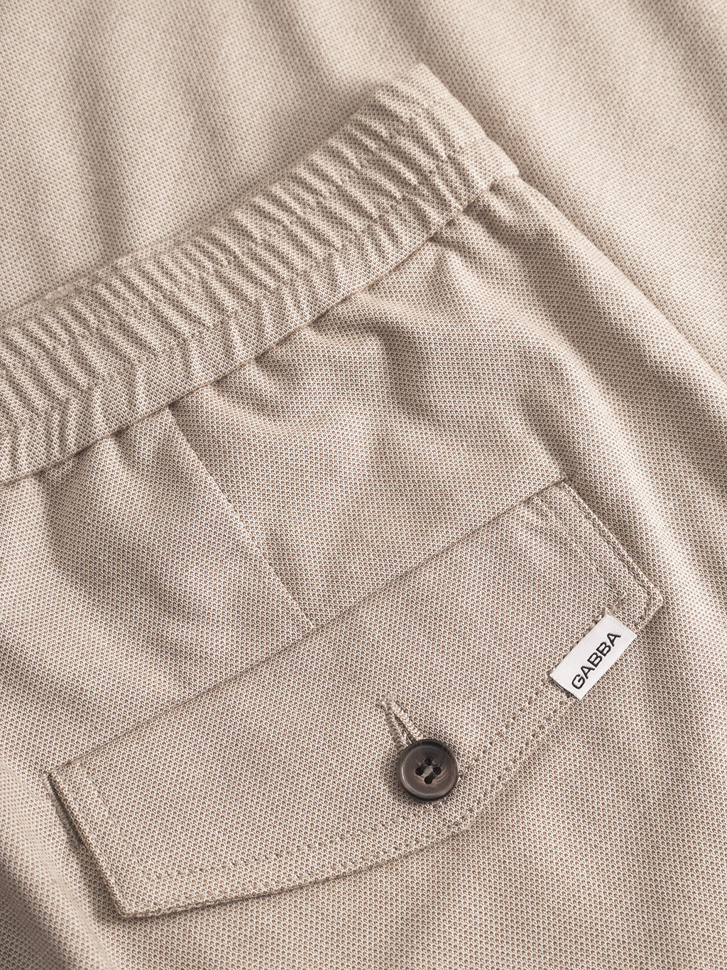 Close-up of the Gabba Monza Button Carre pant in sand. Features an elastic waist, straight relaxed fit, and a buttoned back pocket with a white “GABBA” tag for versatile style.