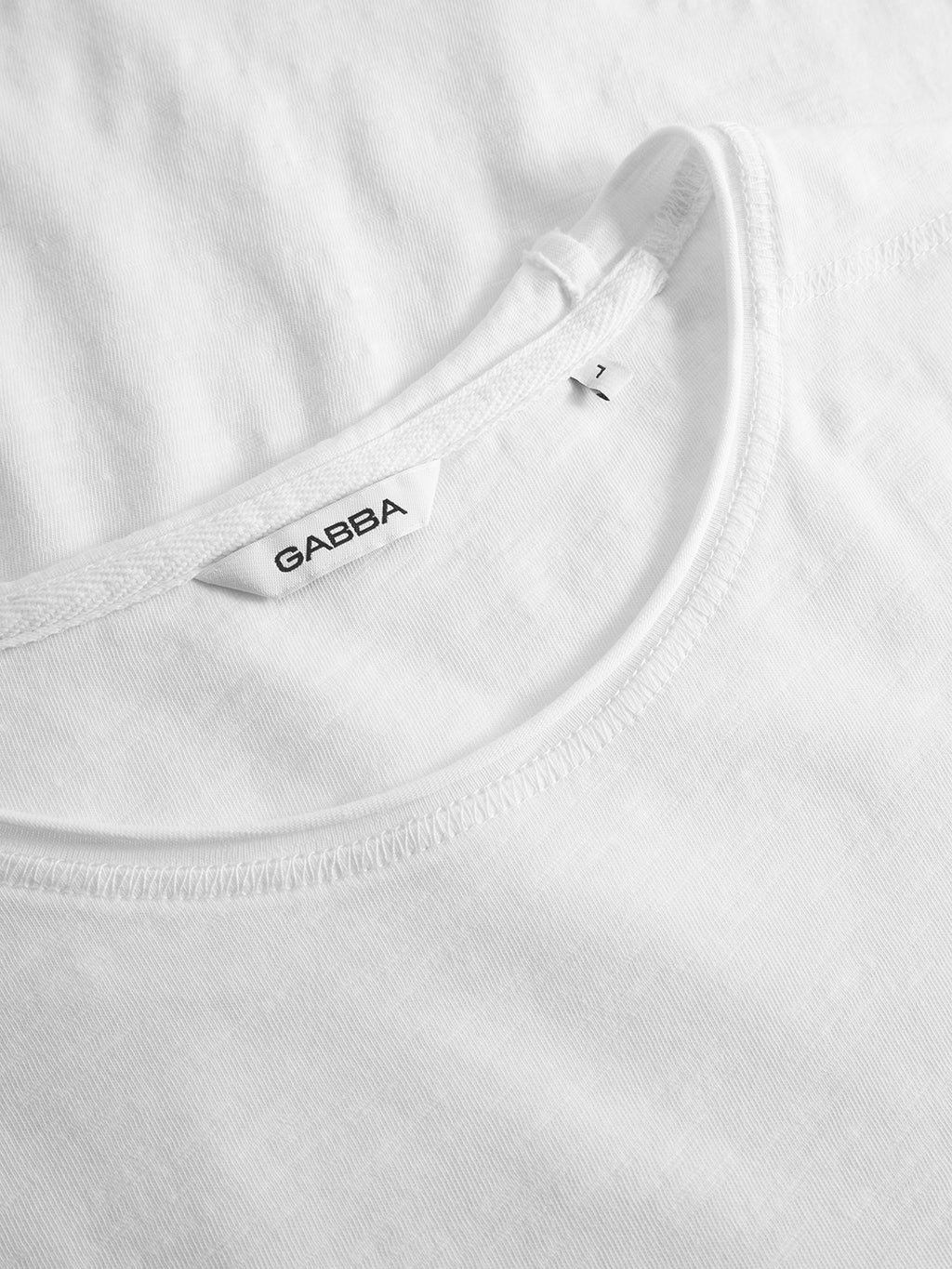 Close-up of the neckline and collar of the GABkostant New SS - White T-shirt by Gabba, showing brand and size labels inside. The 100% cotton slub fabric looks soft, lightweight, and subtly textured.