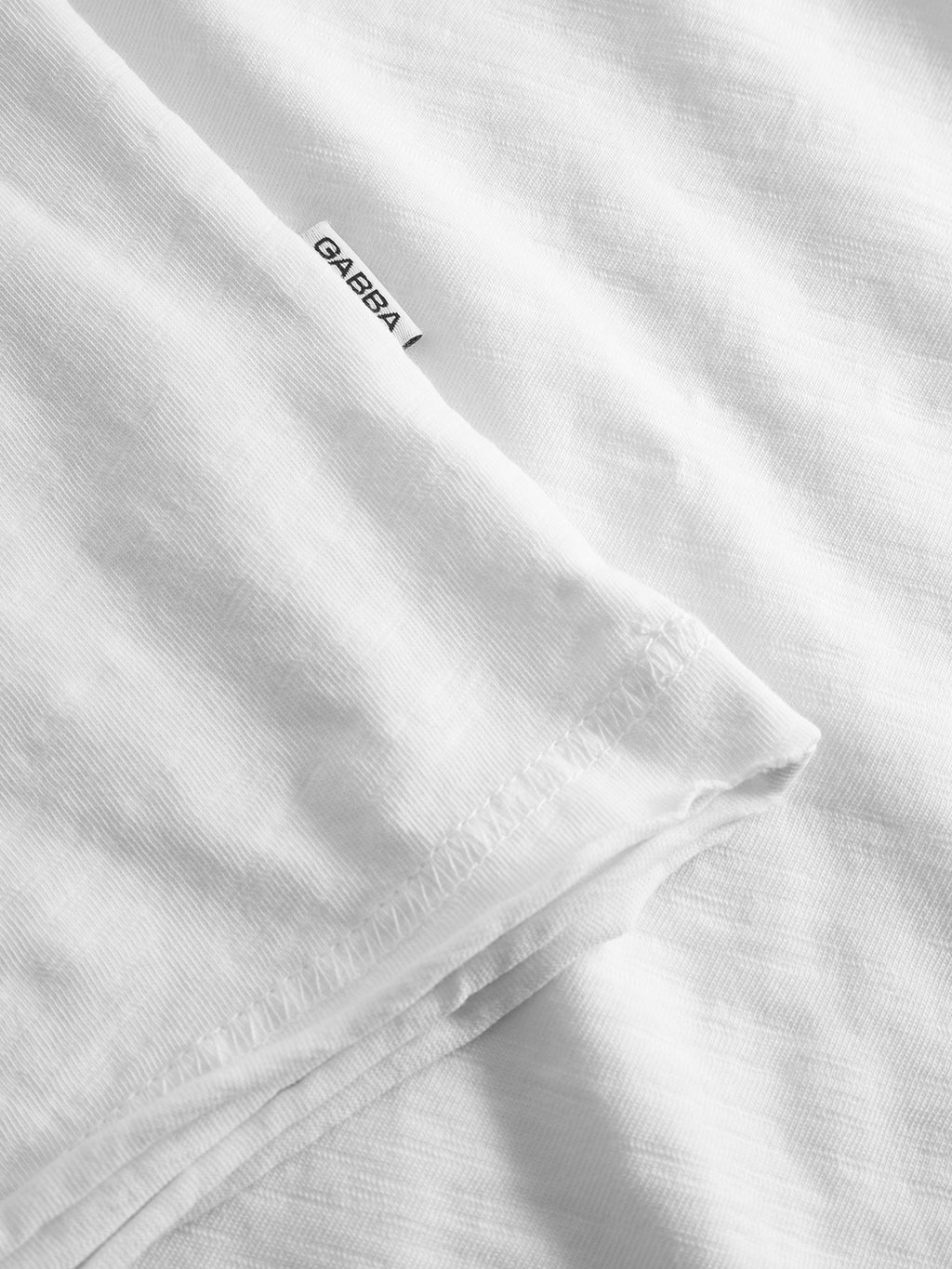Close-up of a white GABkostant New SS t-shirt by Gabba, featuring visible slub fabric texture, detailed stitching, and a small white tag with the Gabba logo in black sewn into the seam.