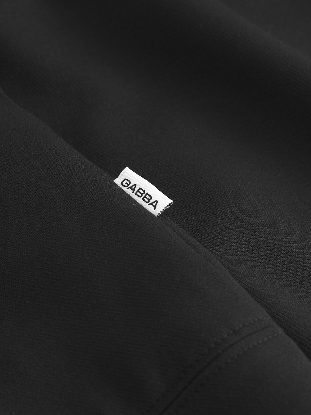 Close-up of the Gabba GABnik Phin GRS - Black jacket, made from recycled polyester, with a small white Gabba tag in black letters partially tucked into a seam.