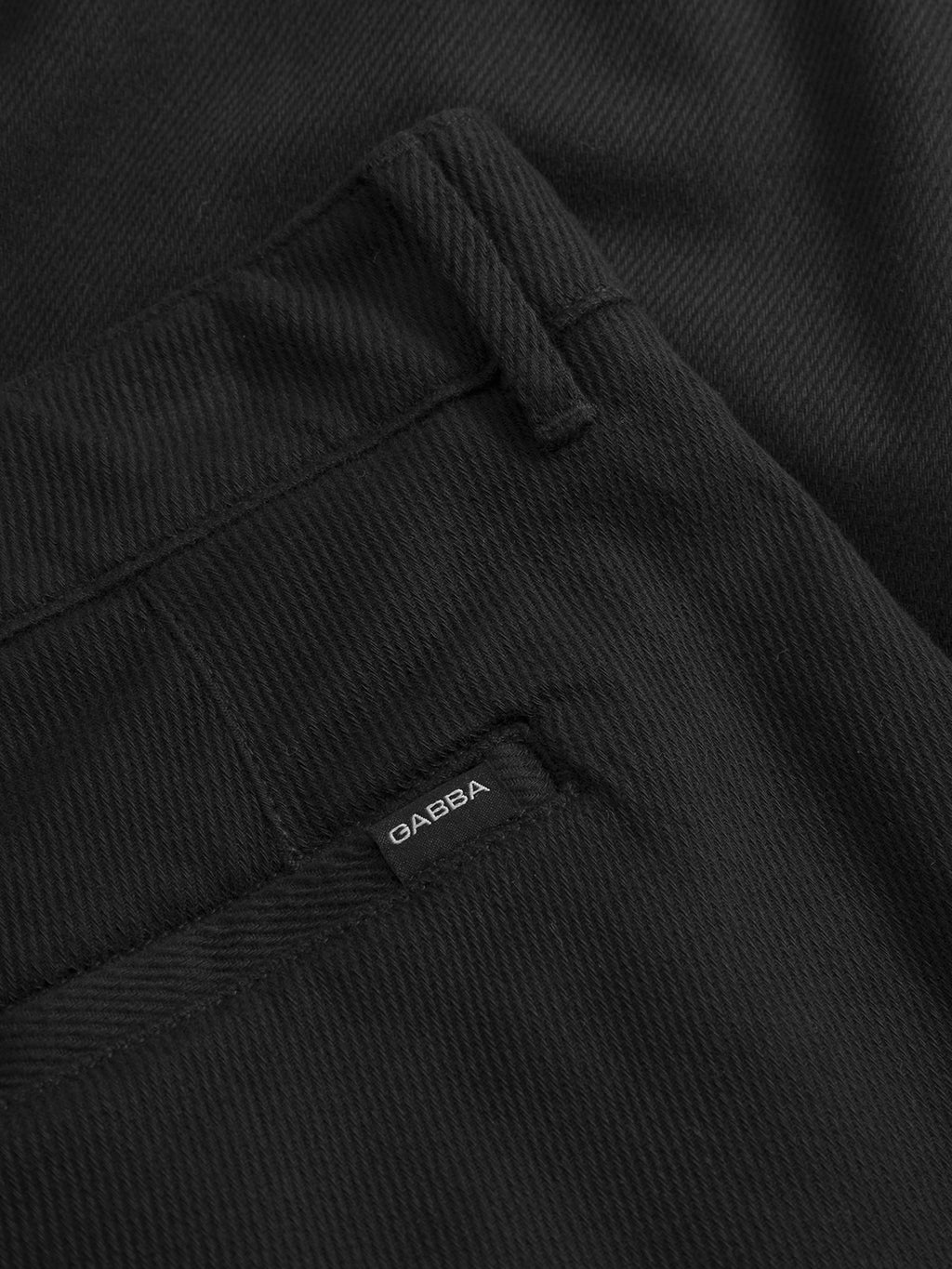 Close-up of the GABJoe‑Pas Vill - Black denim pants by Gabba, designed in a relaxed fit inspired by classic utility trousers. The image highlights a back pocket and a small black fabric label with white “GABBA” text stitched near the pocket. The drapey cotton twill fabric texture and meticulous stitching details are clearly visible.