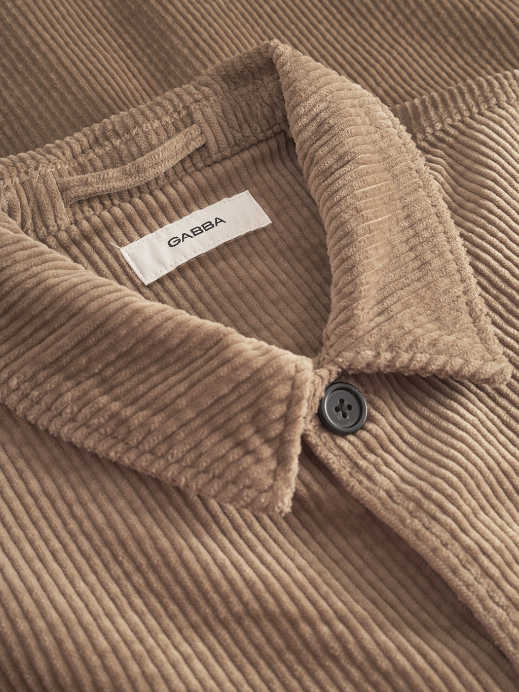 Close-up of the Gabba Libre AX Cord - Sand overshirt featuring a folded collar, black button, and a white label with 