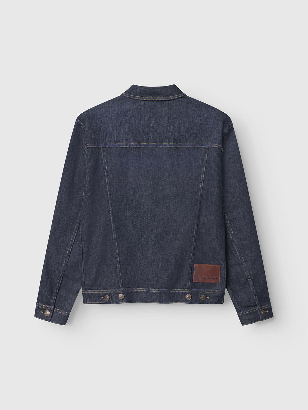 The Luca Self - Dark Blue Denim jacket by Gabba is shown from the back with classic stitching, buttoned cuffs, and a brown leather patch above the hem. Designed sustainably, it’s laid flat on a white background.