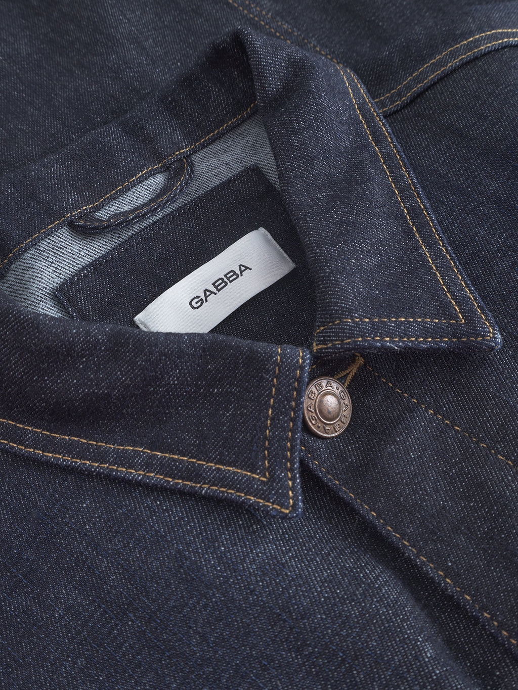 Close-up of the Luca Self - Dark Blue Denim shirt by Gabba, showcasing yellow stitching, a metal button, and a white collar label with the brand name. Pair it with regular-fit jeans for a timeless look.
